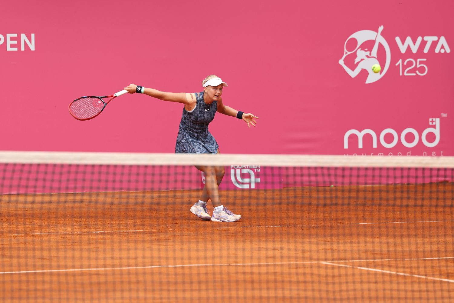 A female tennis player prepares to hit a forehand on a clay court during a WTA 125 tournament, with a bright pink background and court sponsor logos visible.