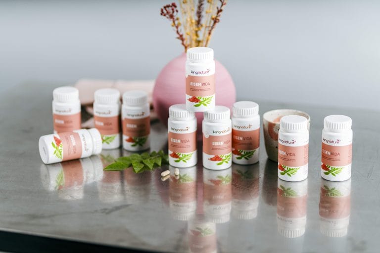 Mehrere Flaschen mit Eisen Vida Nahrungsergänzungsmitteln sind auf einer reflektierenden Oberfläche angeordnet, im Hintergrund steht eine rosa Vase mit getrockneten Pflanzen. Einige Flaschen mit Eisen Vida Eisenpräparaten stehen aufrecht, andere liegen.