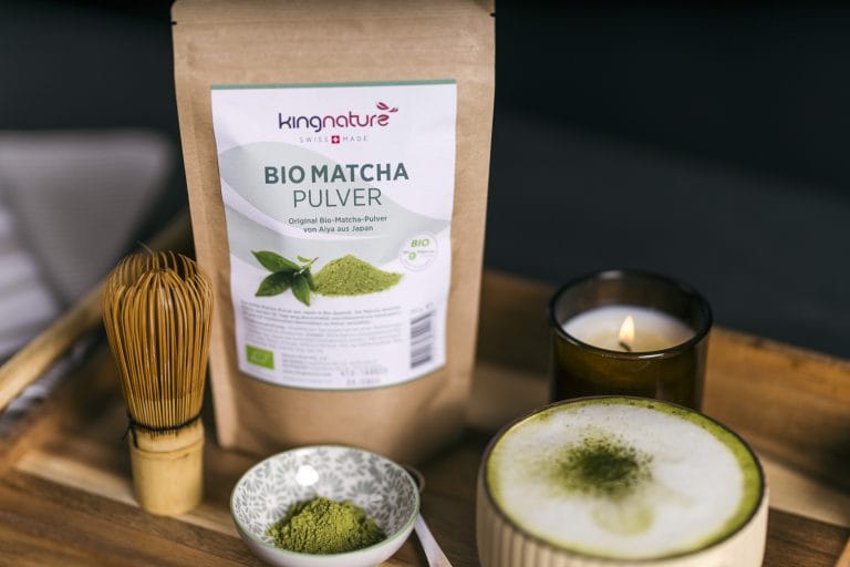 Ein Beutel Kingnature Bio Matcha-Pulver liegt auf einem Holztablett mit einem Bambusbesen, einer Schale mit Bio-Matcha-Pulver, einer brennenden Kerze und einer Tasse kräftigem Matcha-Tee mit grünem Pulver.