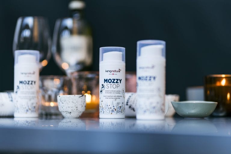 Zwei Flaschen MozzyStop-Insektenschutzmittel werden auf einem reflektierenden Tisch präsentiert, umgeben von kleinen Schüsseln, Tassen und sanft leuchtenden Kerzen in einer warm beleuchteten Innenumgebung.