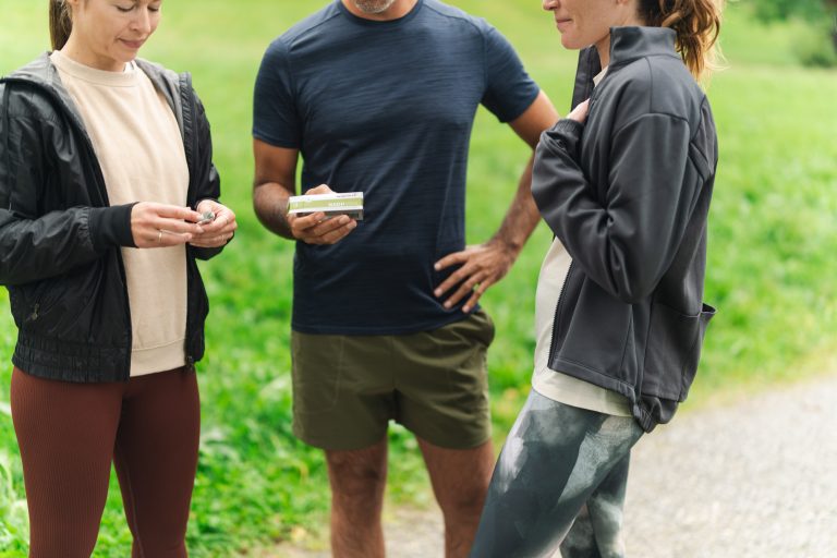Drei Personen in Sportkleidung stehen draußen auf einem Weg mit grünem Gras im Hintergrund. Eine Person hält eine Schachtel, eine andere betrachtet einen Fitnesstracker in ihren Händen. Ihre Gesichter sind nicht vollständig zu sehen.