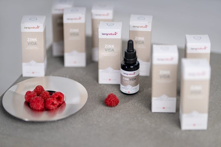 Eine Flasche Zink Vida Zink-Tropfen mit Pipette steht vor mehreren weiß-beigen Kartons mit der Aufschrift „Zink Vida“. Links steht auf einer grauen Fläche ein Teller mit frischen Himbeeren und einer einzelnen Himbeere.