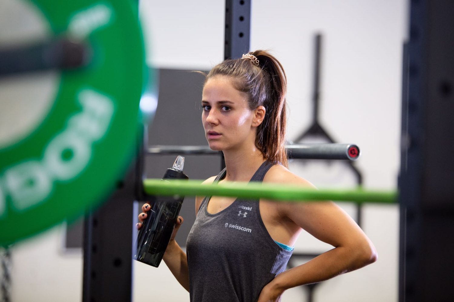 Talina Gantenbein steht in Sportkleidung in einem Fitnessstudio, hält eine Wasserflasche in der Hand und blickt in die Kamera. Im Hintergrund sind Fitnessgeräte und Gewichte zu sehen.