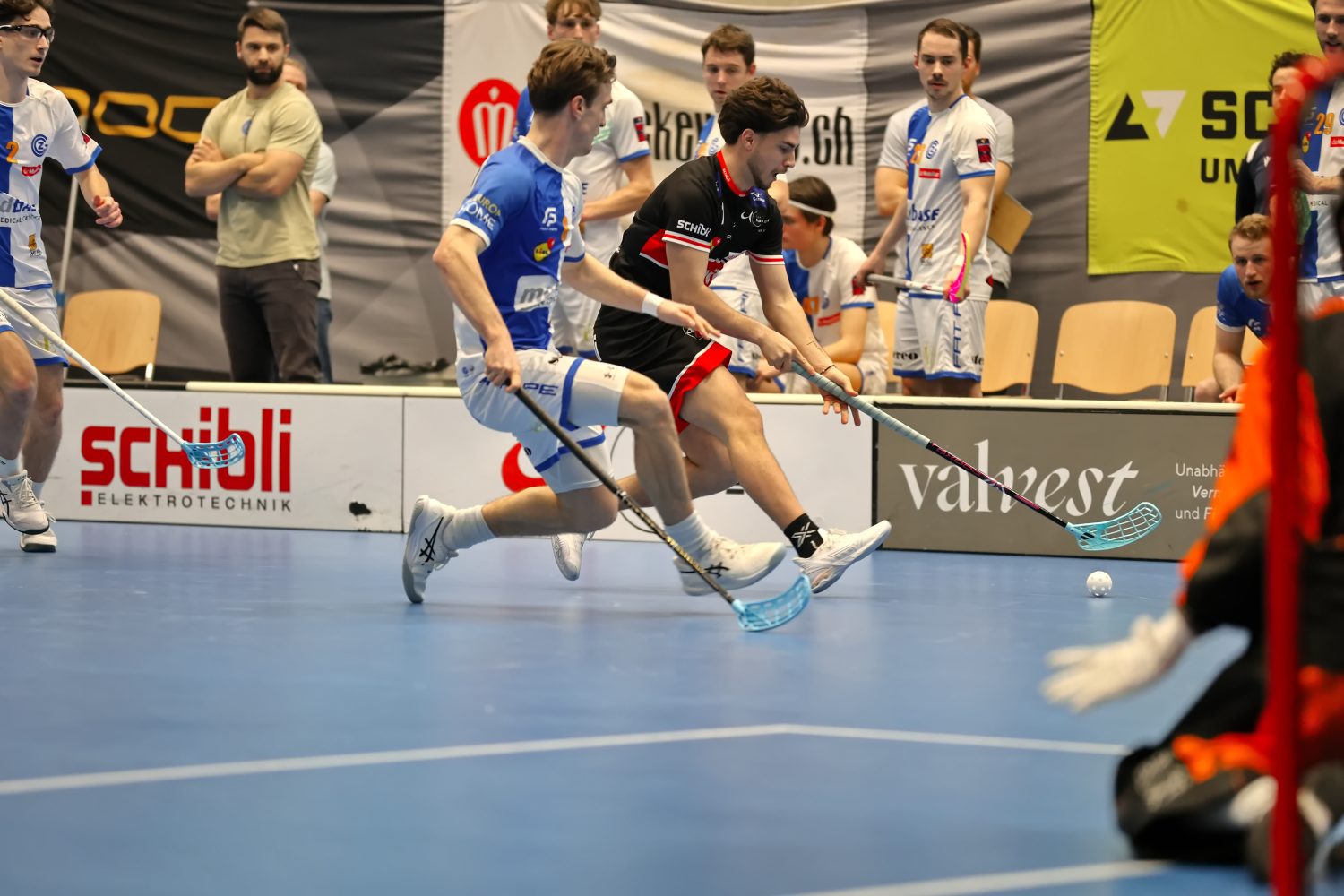 Zwei männliche Unihockeyspieler, darunter Josia Pfister in schwarzer Uniform, kämpfen auf einem Hallenplatz um den Ball. Im Hintergrund sind weitere Spieler und Zuschauer zu sehen.