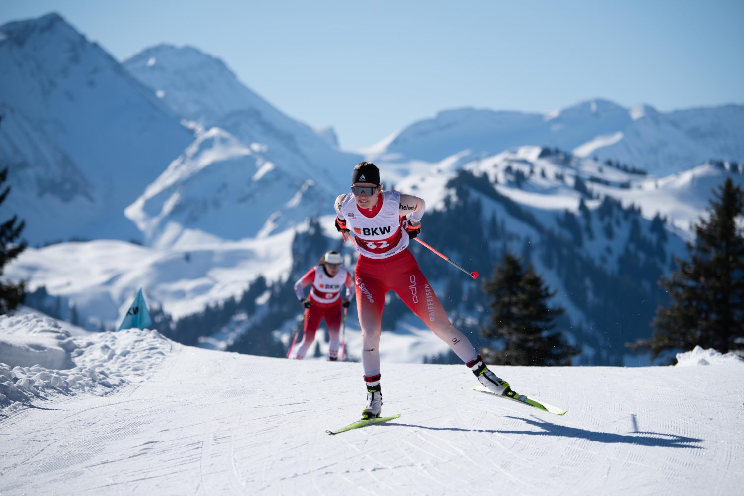 Zwei Langläufer liefern sich unter klarem, blauem Himmel ein Rennen auf einer verschneiten Bergloipe. Ramona Schöpfer führt in einer rot-weißen Uniform und gleitet vor der atemberaubenden Kulisse schneebedeckter Gipfel und Tannenbäume voran.