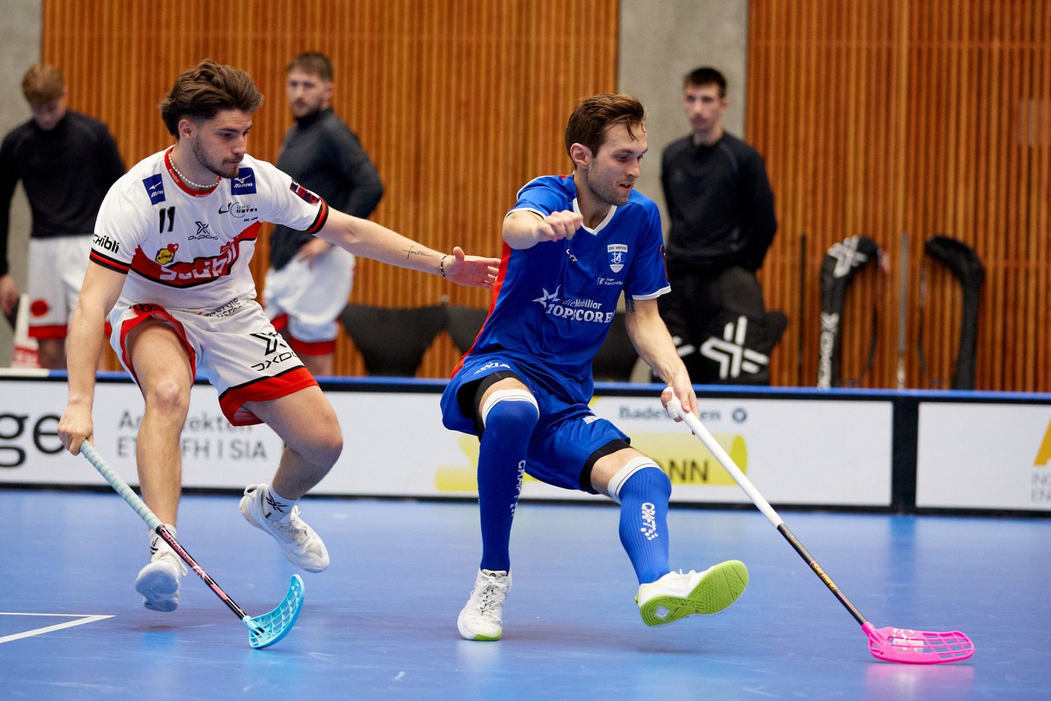 Zwei männliche Unihockeyspieler kämpfen um den Ball. Josia Pfister, in Weiß, streckt die Hand nach dem Spieler in Blau aus, der mit dem Ball nach vorne läuft. Im Hintergrund sind Zuschauer und Unihockeyausrüstung zu sehen.