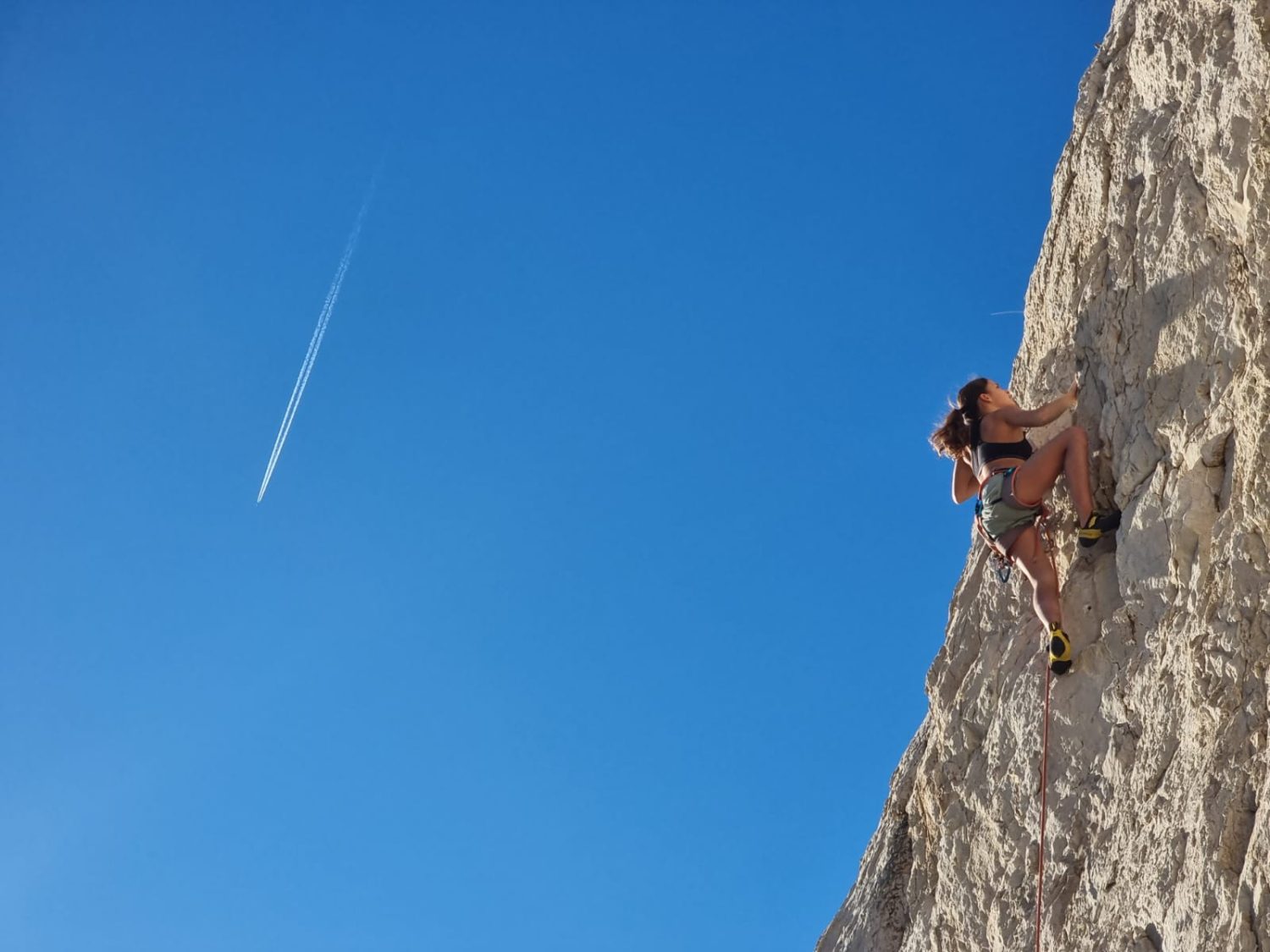 Valentina von Gunten erklimmt, ausgerüstet mit Kletterausrüstung, unter strahlend blauem Himmel eine steile Felswand. Links in der Ferne hinterlässt ein Flugzeug einen sichtbaren Kondensstreifen.