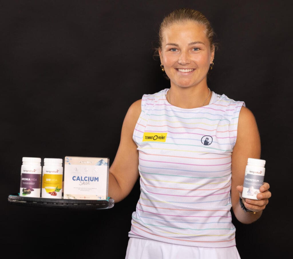 Eine lächelnde Frau in einem gestreiften ärmellosen Hemd hält vor einem schlichten schwarzen Hintergrund ein Tablett mit verschiedenen Nahrungsergänzungsmittelflaschen und einer Schachtel sowie eine weitere Flasche in der anderen Hand und betont damit die tägliche Ernährung und das Wohlbefinden.