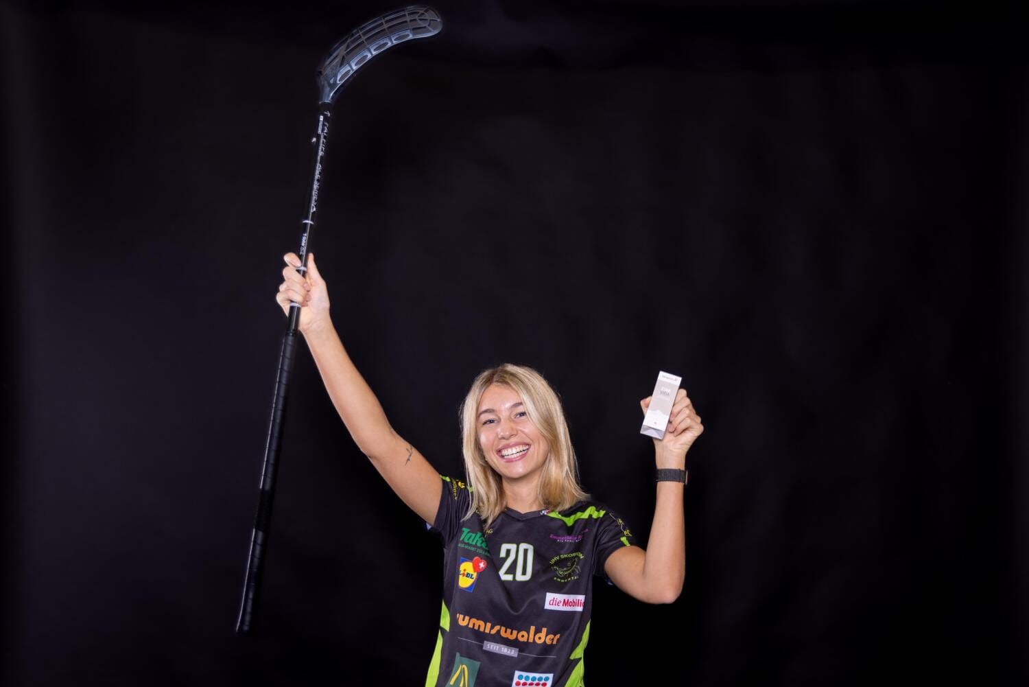 Leonie Soraya Mai, lächelnd in einem Sporttrikot, hält in der einen Hand einen Unihockeyschläger und in der anderen eine kleine weiße Schachtel und posiert aufgeregt vor einem schlichten schwarzen Hintergrund.