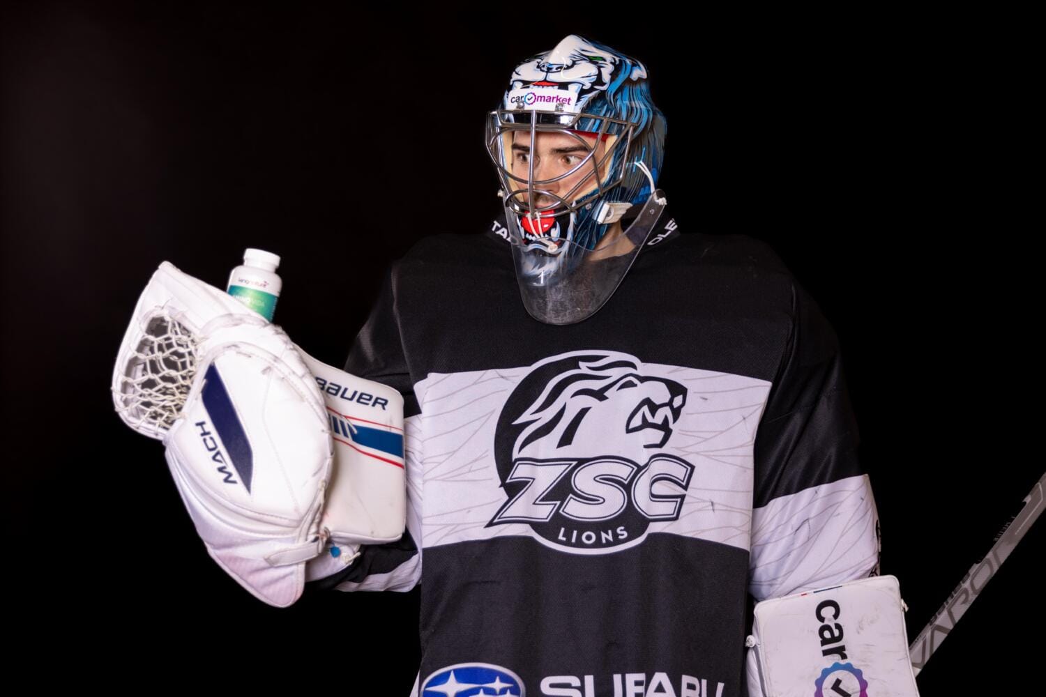 Hockey-Torwart Jeffrey Meier trägt ein schwarzes ZSC Lions-Trikot und einen blau-weissen Helm und hält vor einem schwarzen Hintergrund einen Torwarthandschuh und eine Wasserflasche.