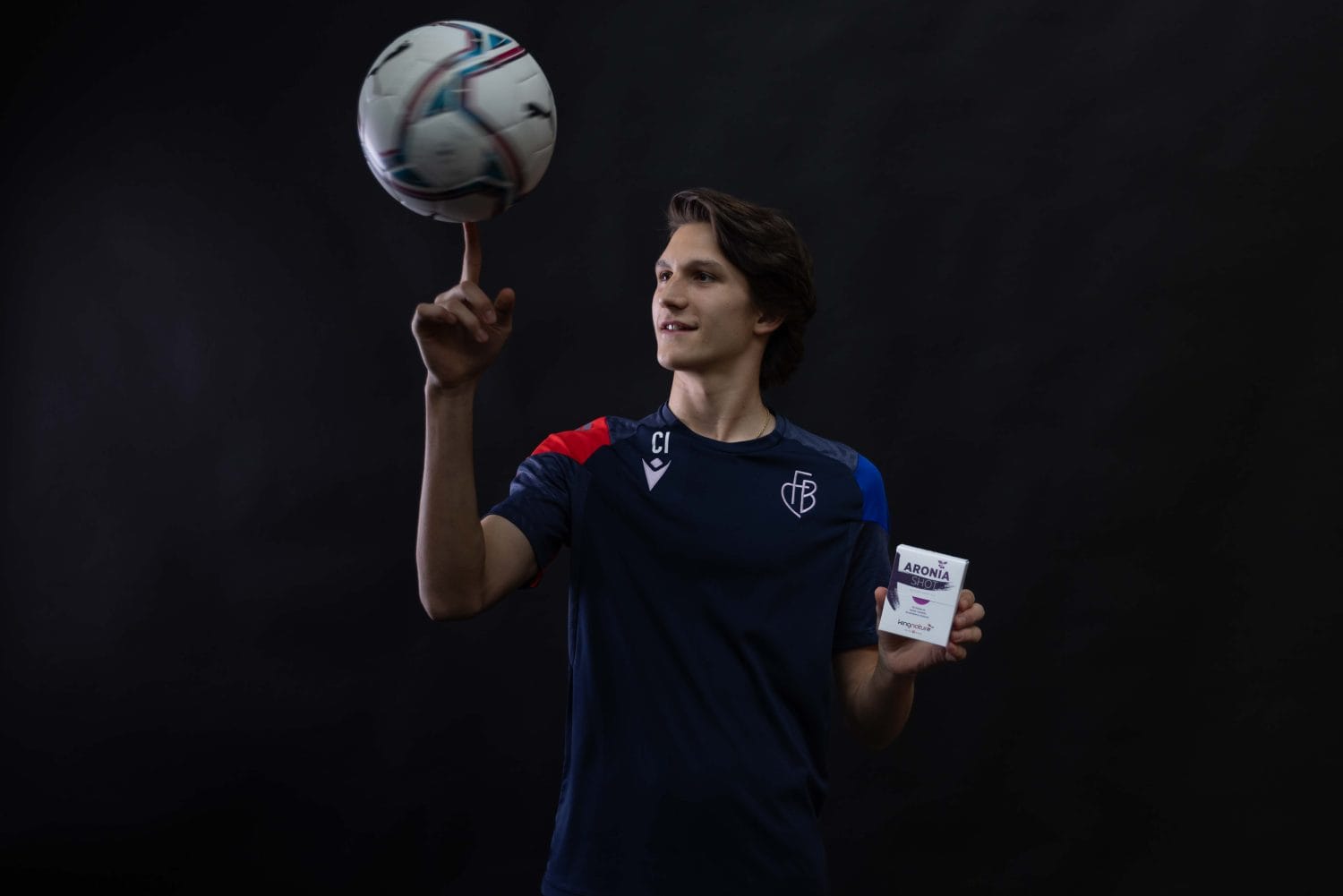 Vor einem dunklen Hintergrund lässt ein junger Mann in einem dunklen Sporttrikot mit einer Hand gekonnt einen Fußball auf seinem Finger kreisen und hält in der anderen einen kleinen weißen Behälter.