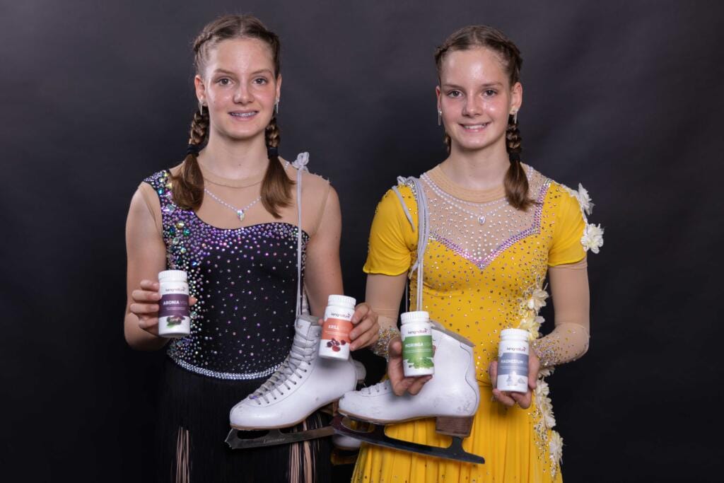 Selina Arnold und Alessia Arnold, zwei Mädchen mit geflochtenen Haaren in glitzernden Eiskunstlaufkleidern (eines schwarz, eines gelb), halten Schlittschuhe und präsentieren Flaschen mit Nahrungsergänzungsmitteln vor einem schlichten dunklen Hintergrund.