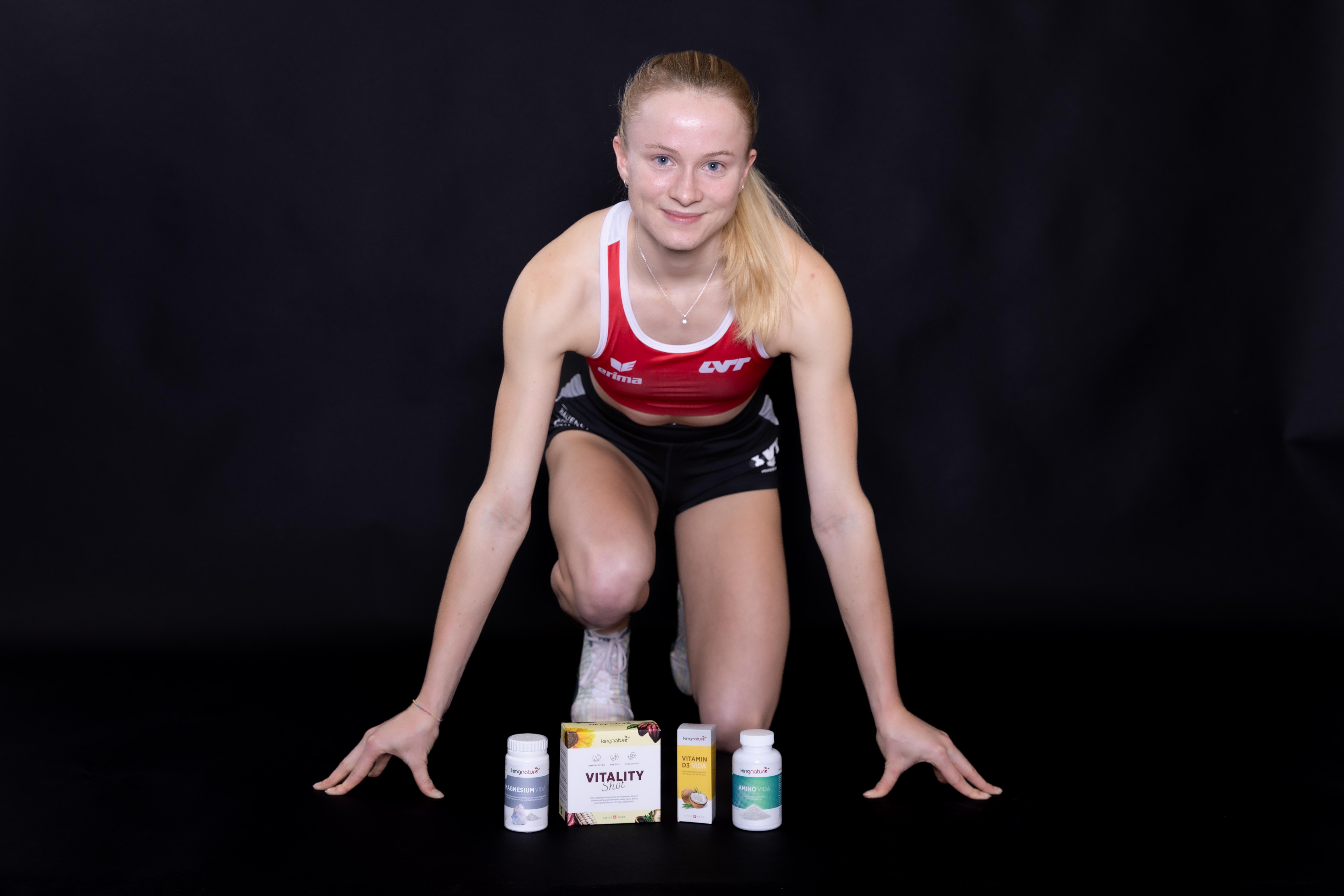 Linda Bichsel, eine Sportlerin in Sportkleidung, kniet lächelnd in Startposition vor schwarzem Hintergrund, vor ihr sind vier Behälter mit Nahrungsergänzungsmitteln ausgestellt.