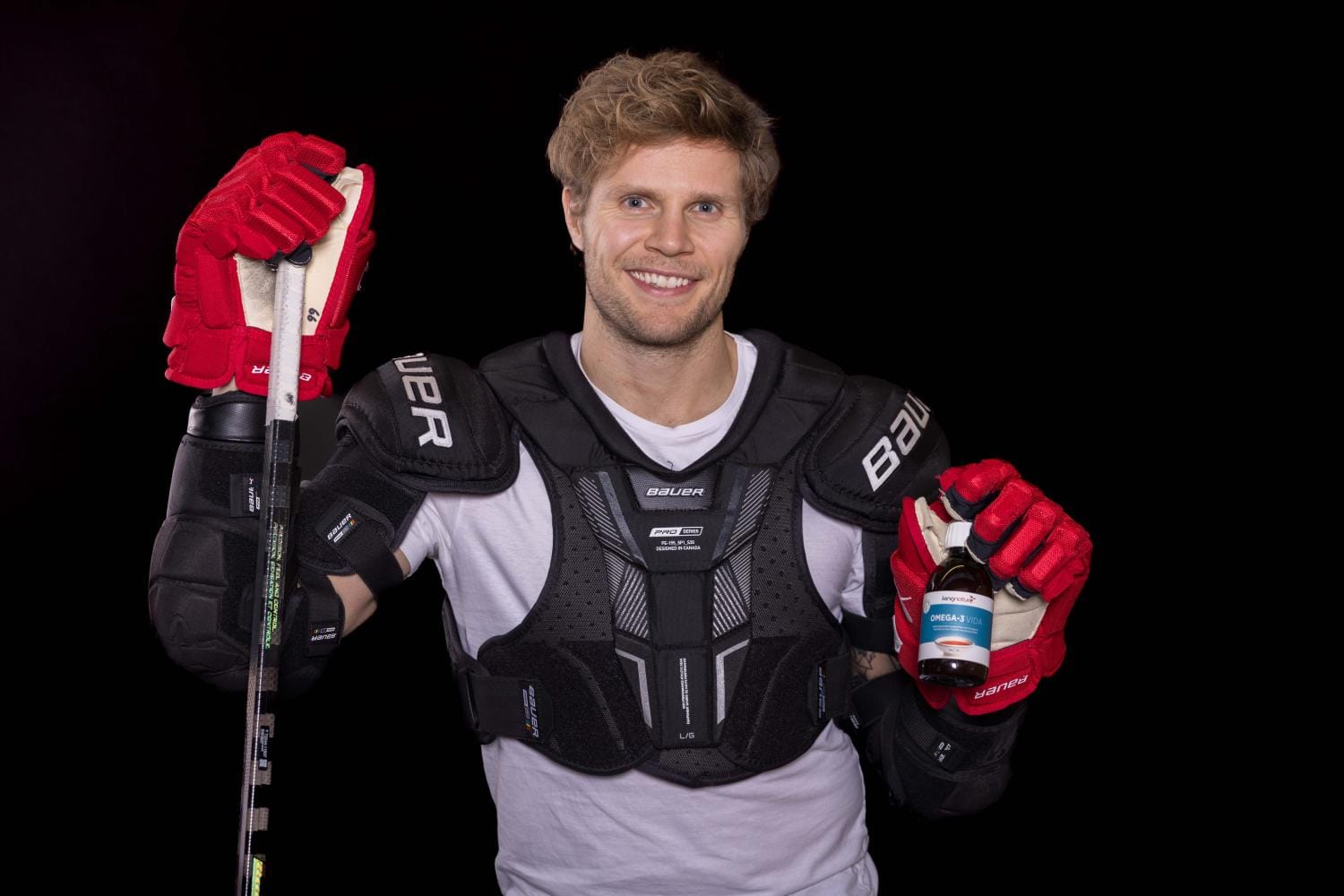 Ein lächelnder Hockeyspieler, Tim Grossniklaus, in schwarzer Schutzausrüstung und roten Handschuhen, hält einen Hockeyschläger in der einen Hand und eine kleine Flasche in der anderen und steht vor einem schwarzen Hintergrund.