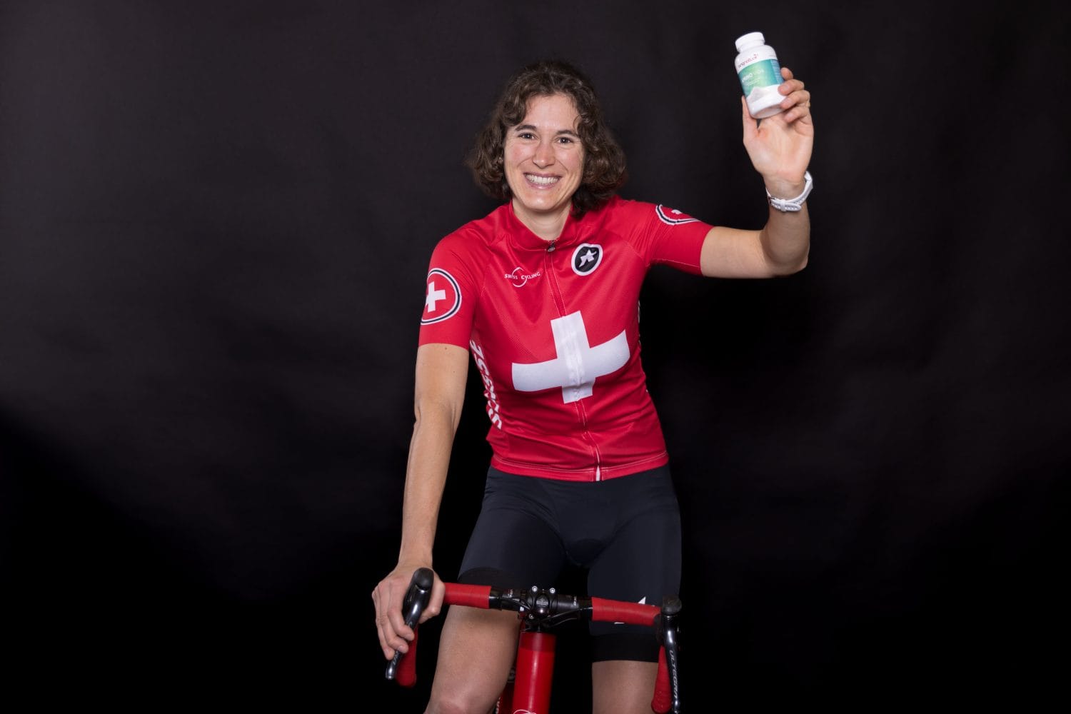 Eine lächelnde Radfahrerin, Franziska Matile-Dörig, in einem roten Trikot mit einem weißen Kreuz, sitzt auf einem Heimtrainer und hält vor einem schwarzen Hintergrund eine kleine Flasche hoch.