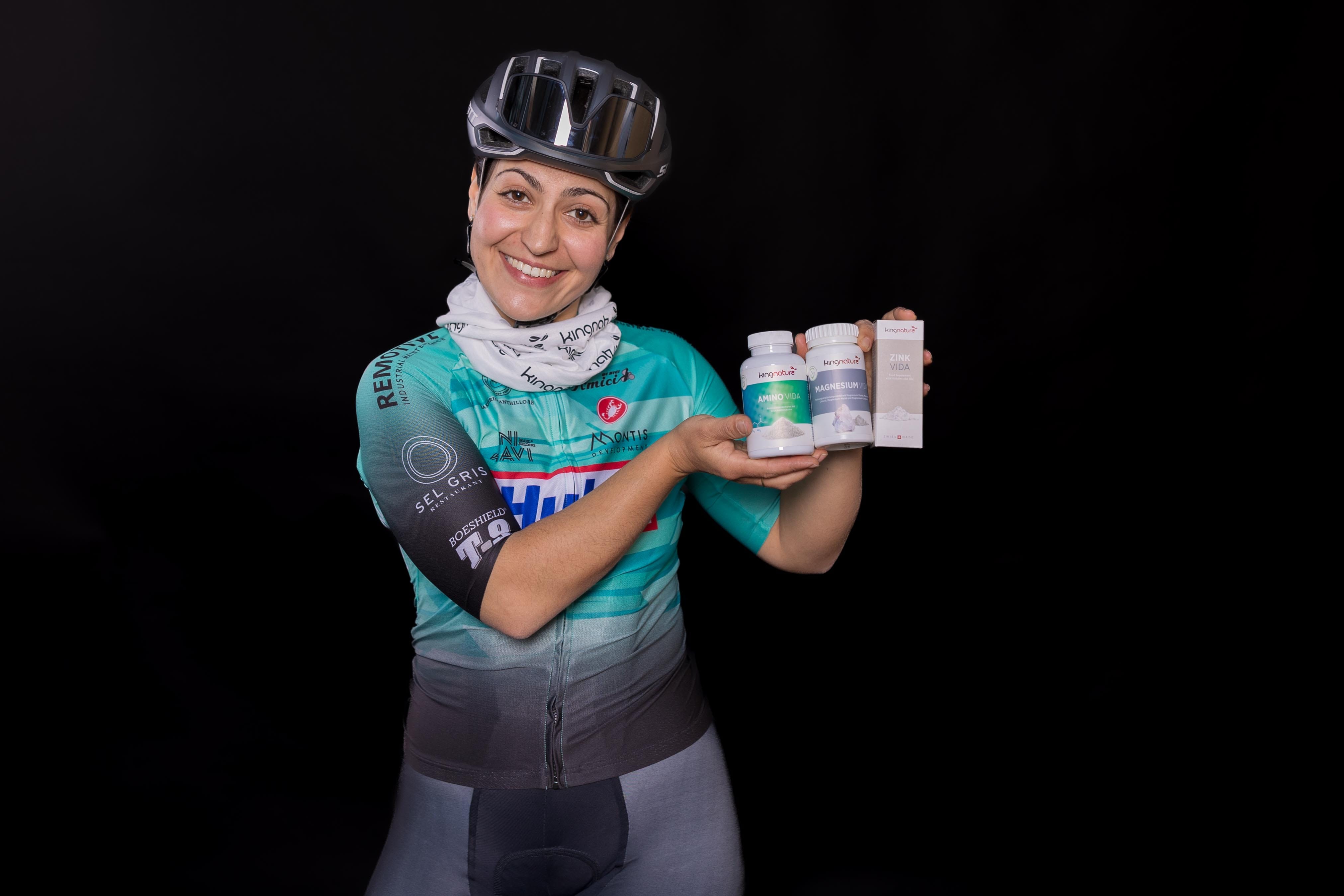 Zina Barhoumi, eine Radfahrerin mit Helm und blaugrünem Trikot, lächelt, während sie vor einem schwarzen Hintergrund drei Flaschen und zwei Schachteln mit Nahrungsergänzungsmitteln hochhält.