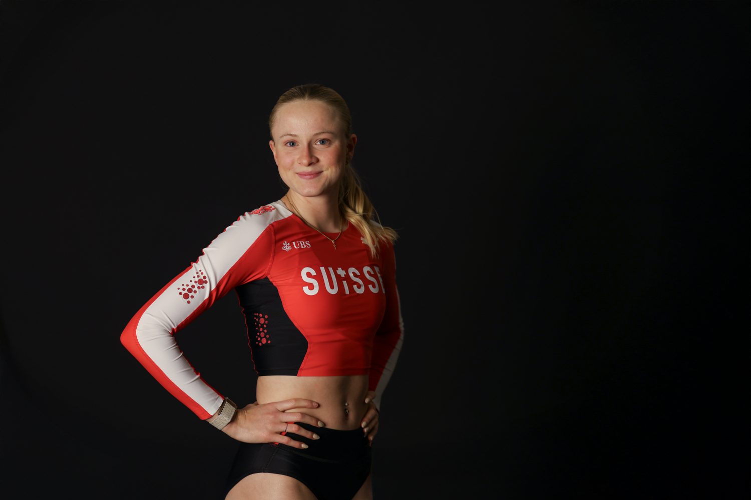 Linda Bichsel, eine Sportlerin, steht selbstbewusst mit den Händen in den Hüften und trägt ein rot-weißes langärmliges bauchfreies Top mit SUI-Schriftzug und schwarze Shorts vor einem einfarbig schwarzen Hintergrund.
