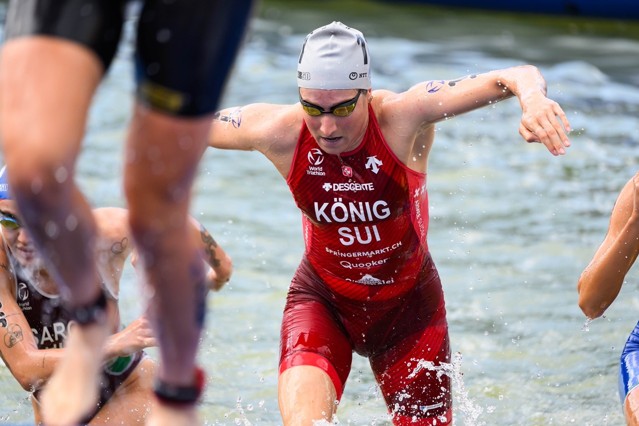 Eine Triathletin in rotem Anzug und grauer Badekappe, Alissa König, steigt mit ausgestreckten Armen aus dem Wasser und nimmt an einem Rennen teil. Um sie herum sind andere Athleten zu sehen, mit Spritzern und Wassertropfen in Bewegung.