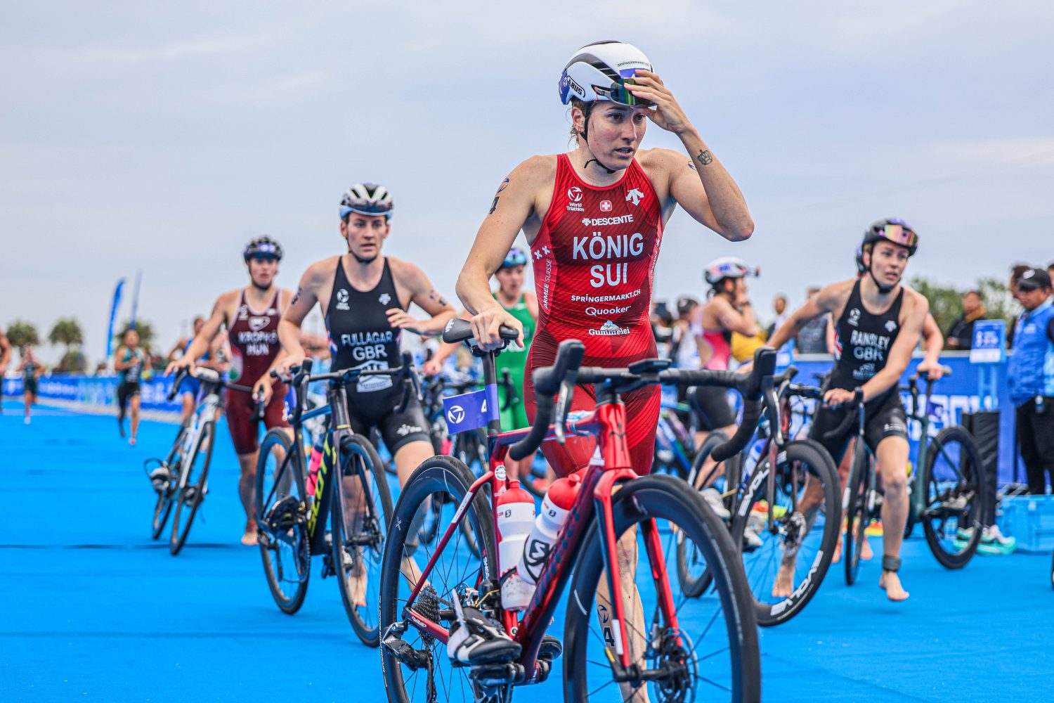 Triathleten in Rennmontur und mit Helmen schieben ihre Räder durch eine blaue Wechselzone und bereiten sich auf die Radstrecke vor. Angeführt wird die Gruppe von Alissa König, die einen roten Anzug mit der Aufschrift „König SUI“ trägt. Konzentriert und bereit für die nächste Herausforderung.