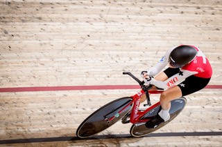 Eine Radfahrerin, Franziska Matile-Dörig, trägt eine rot-weiße Uniform und einen schwarzen Helm und fährt ein rotes Rennrad auf einer Indoor-Velodrombahn, aufgenommen von oben.