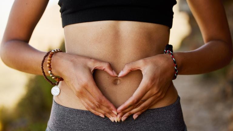 Eine Person in einem schwarzen Oberteil und einer grauen Hose formt mit ihren Händen auf dem nackten Bauch ein Herz und präsentiert dabei stylische Armbänder an beiden Handgelenken.