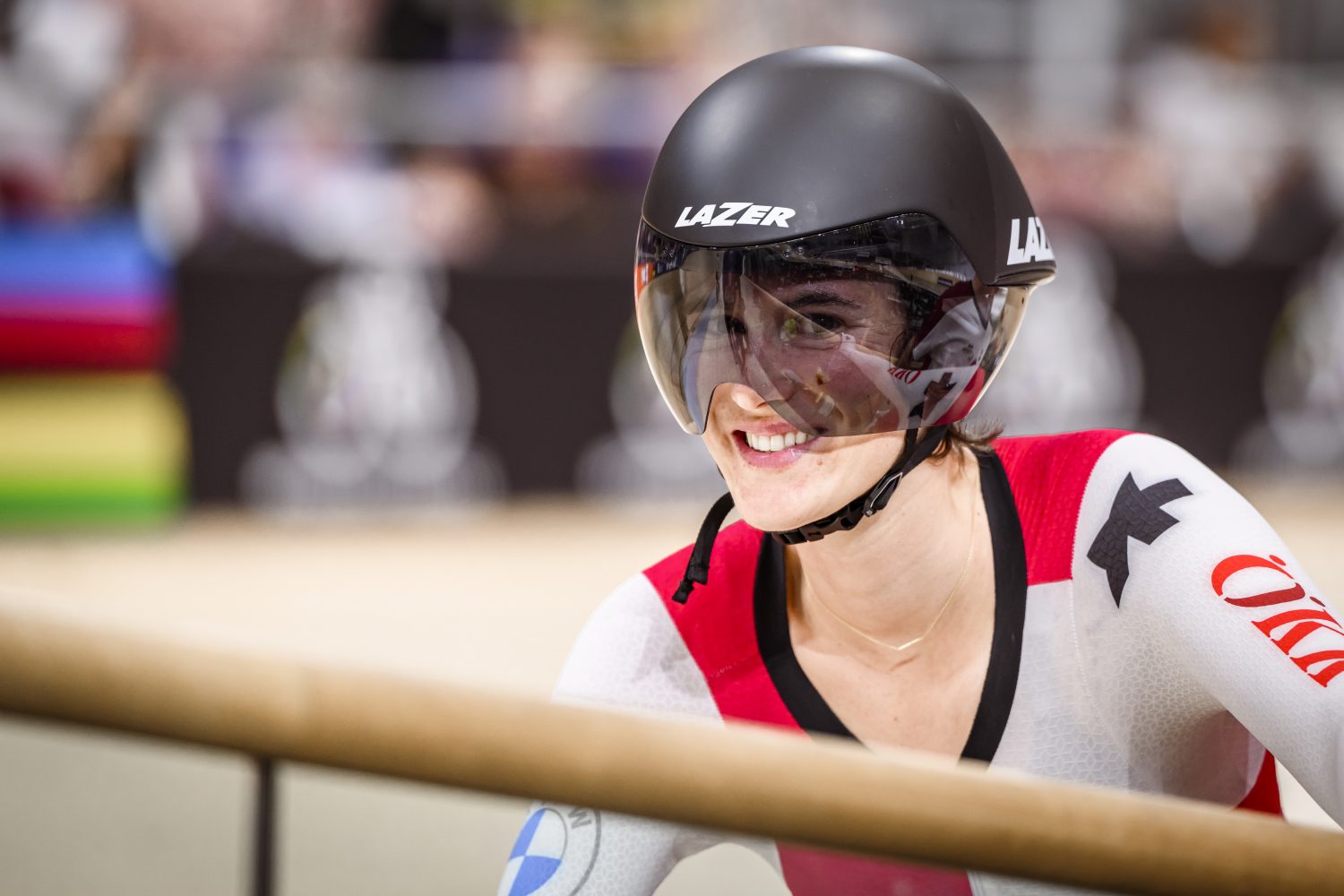 A smiling cyclist wearing a black helmet and white racing suit with red and black accents stands in an indoor velodrome.