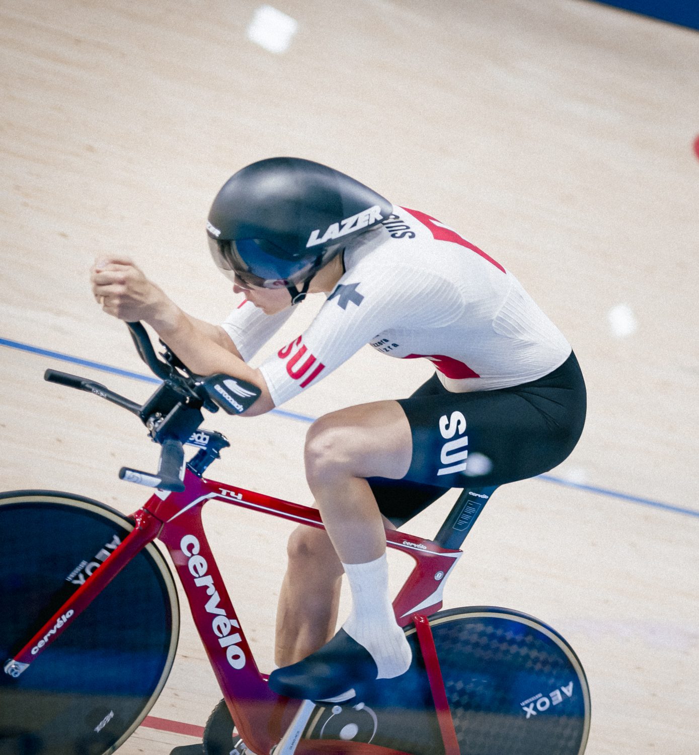 A cyclist wearing a white and red Swiss uniform races on a track bike at the Paris 2024 Paralympic Games, with the Paralympic symbol and PARIS 2024 visible in the background.