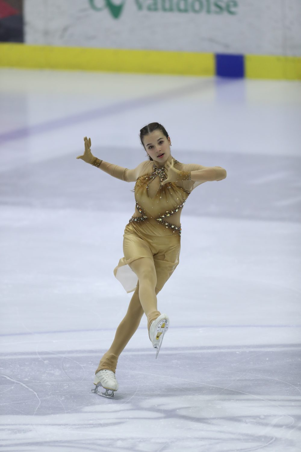 Giada Angelina Molinari tritt in einem goldenen Kostüm auf einer Eisbahn auf, balanciert auf einem Bein mit ausgestreckten Armen und angewinkeltem Bein vor einem Banner der Vaudoise.