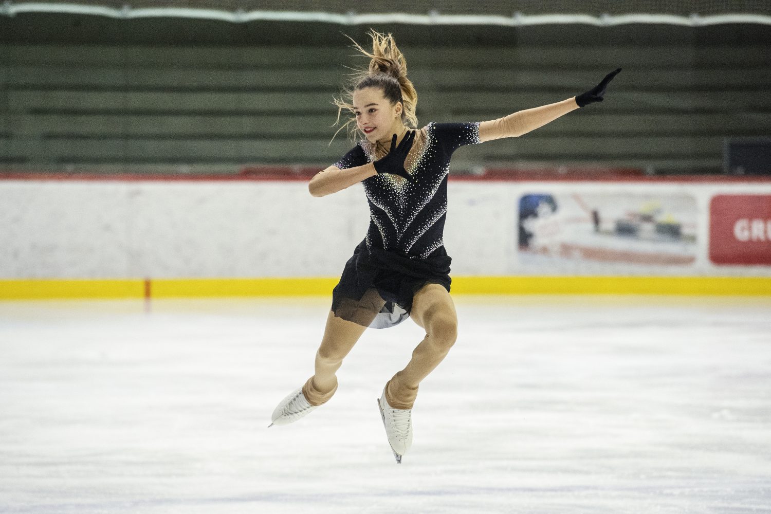 Giada Angelina Molinari führt in einem glitzernden schwarzen Kleid und hellbraunen Strumpfhosen einen Sprung auf einer Eishalle aus, mit angewinkeltem Bein, ausgestreckten Armen und wehendem Haar.