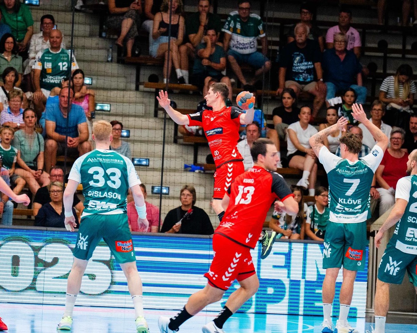 A handball player in a red uniform jumps to shoot the ball while surrounded by opposing players in green uniforms, with a crowd of spectators watching from the stands.