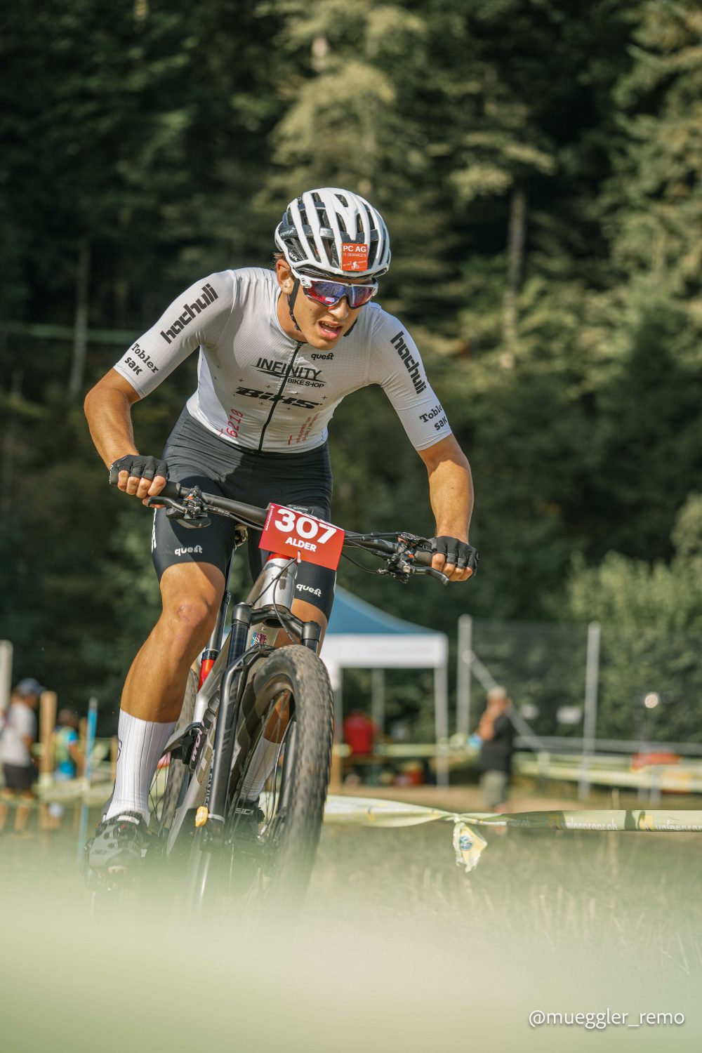Ein Radfahrer mit weißem Helm und Sonnenbrille nimmt an einem Wettkampf auf einem Mountainbike mit der Startnummer 307 teil, umgeben von üppigem Grün und jubelnden Zuschauern im Hintergrund.