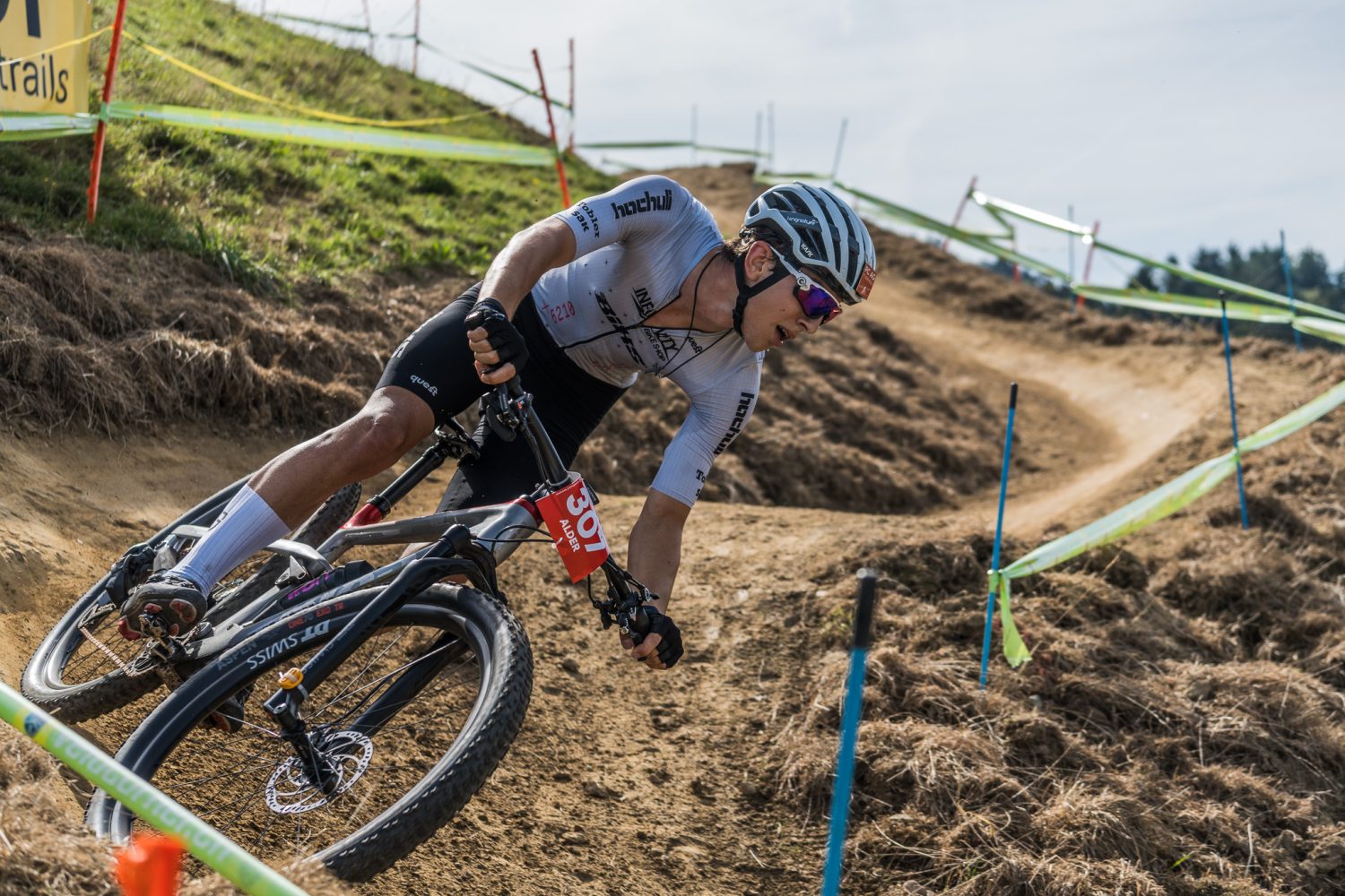 Ein Radfahrer mit Helm und Sonnenbrille rast mit einem Mountainbike um die Kurve eines Feldwegs und legt sich dabei in die Kurve, die an einem grasbewachsenen Hang entlangführt, der mit blauen und gelben Streckenflaggen markiert ist.