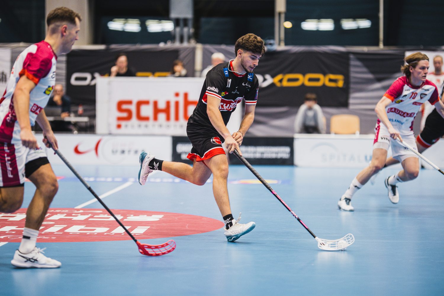 Drei Unihockeyspieler in Aktion auf einem Hallenplatz, darunter Josi Pfister in Weiß und Pink, alle konzentriert auf den Ball. Im Hintergrund sind Zuschauer und Banner zu sehen.