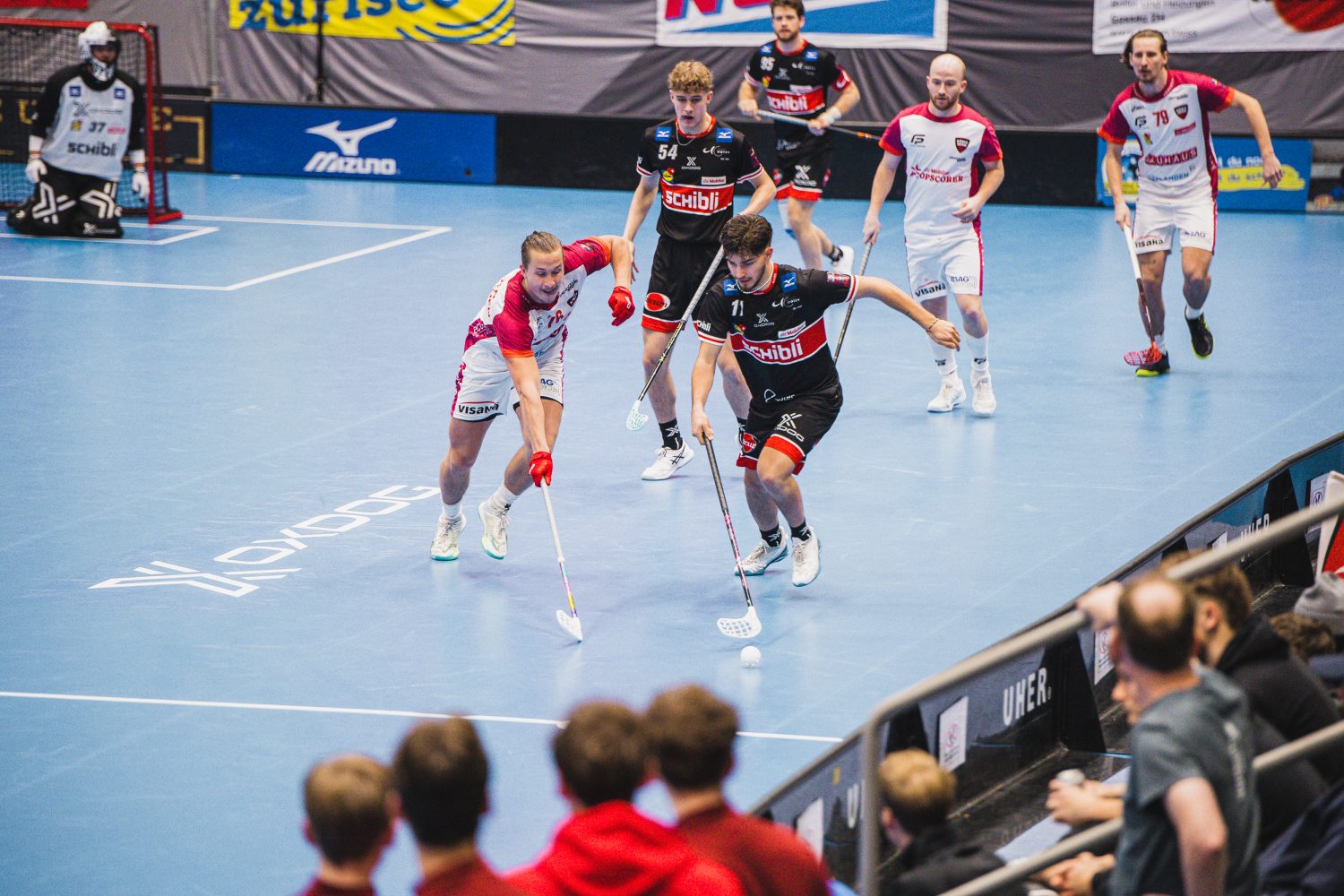 Zwei Unihockey-Teams treten in einem rasanten Match auf einem blauen Hallenplatz gegeneinander an. Spieler in weiß-schwarzen Uniformen – darunter auch Josia Pfister – jagen dem Ball hinterher, während die Zuschauer von der Tribüne aus zuschauen.