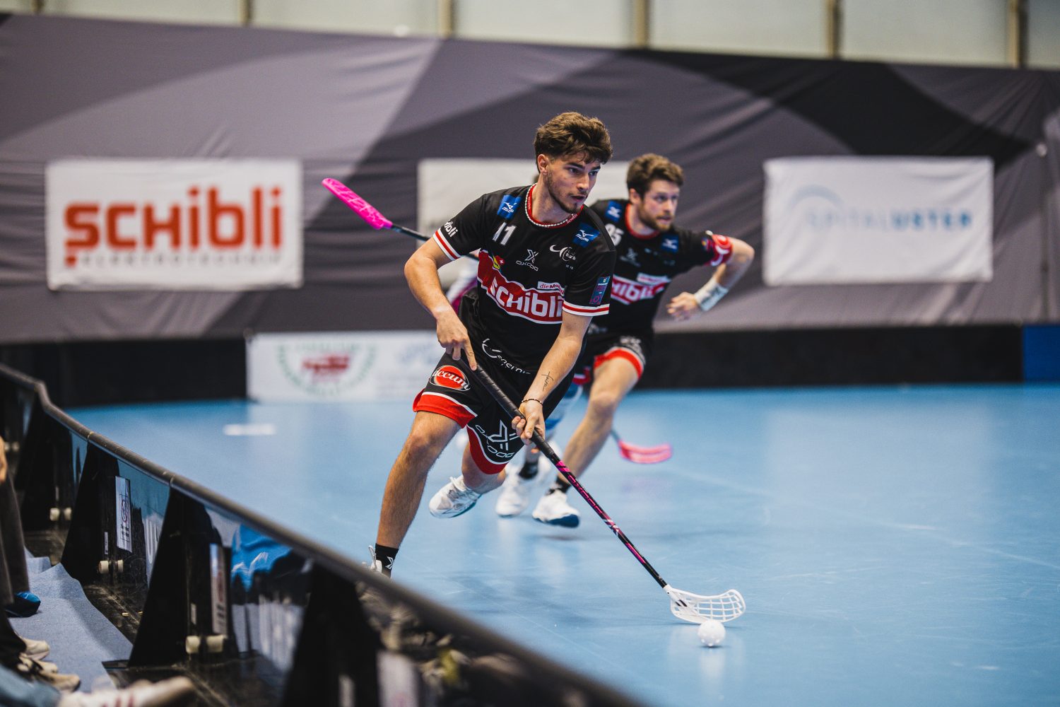 Zwei Unihockeyspieler, darunter Josi Pfister in schwarzer Uniform, treten in einer Halle gegeneinander an. Der führende Spieler kontrolliert den Ball mit seinem Schläger, während der zweite Spieler dicht hinterherläuft. An den Wänden sind Werbebanner zu sehen.