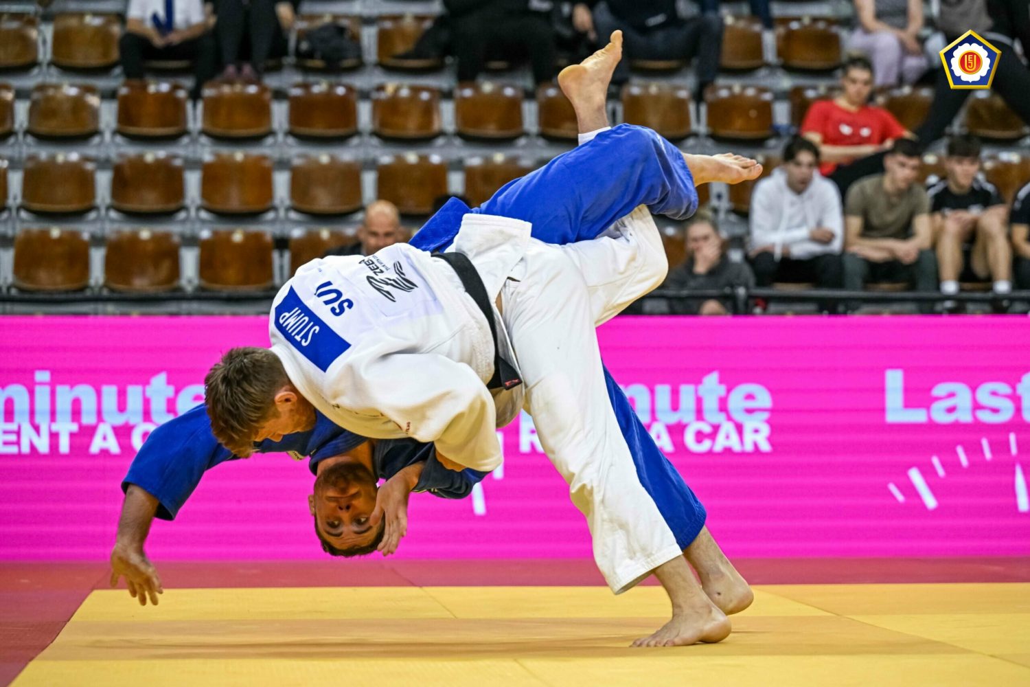 Two judo athletes compete on a mat, with one throwing the other over his shoulder. The competitor in white has SUI on his back, while the competitor in blue is mid-air. Spectators watch from the stands.