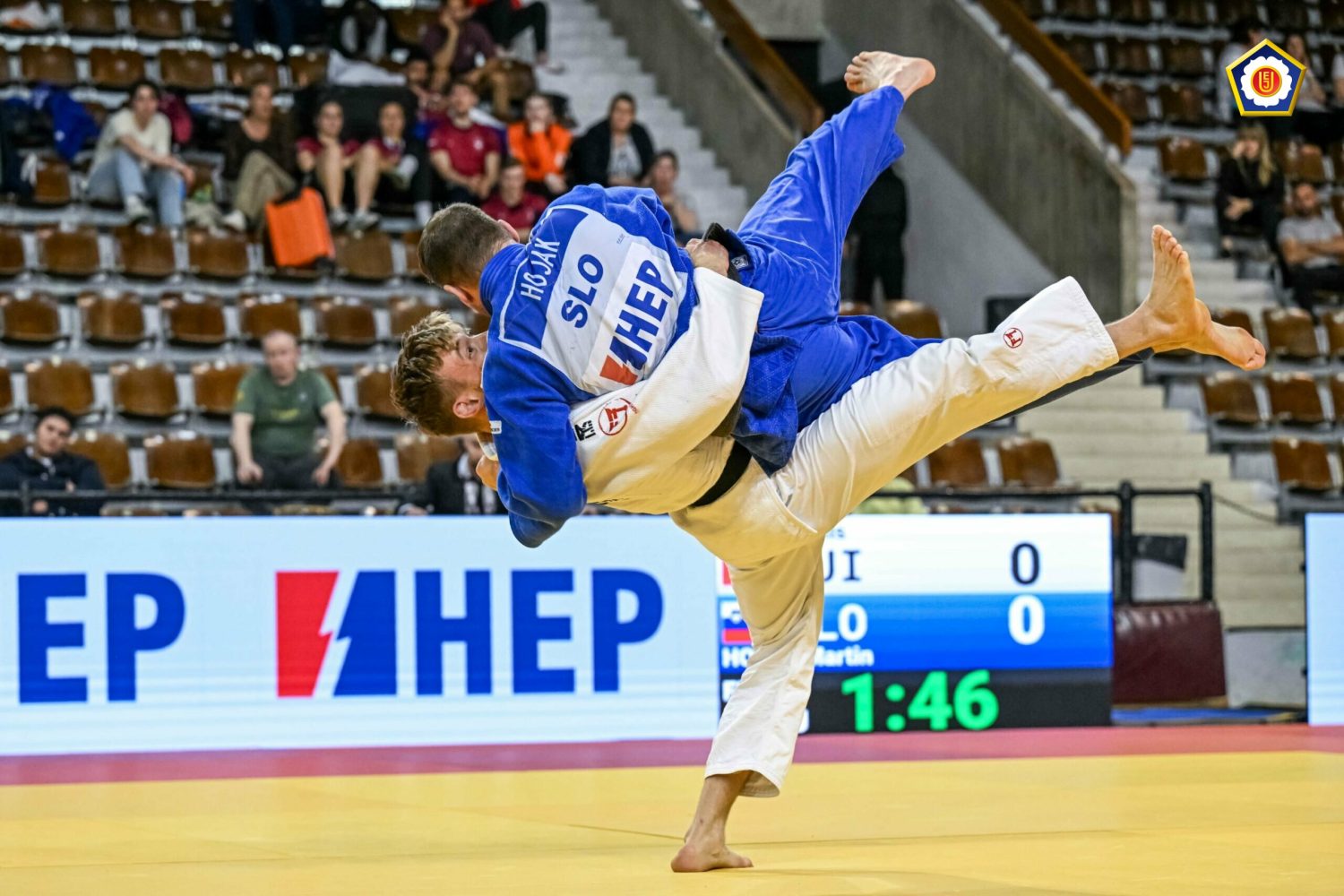 Two judo athletes compete on a mat; one in a blue uniform throws the other in white. Spectators watch from the stands, and a scoreboard is visible in the background.