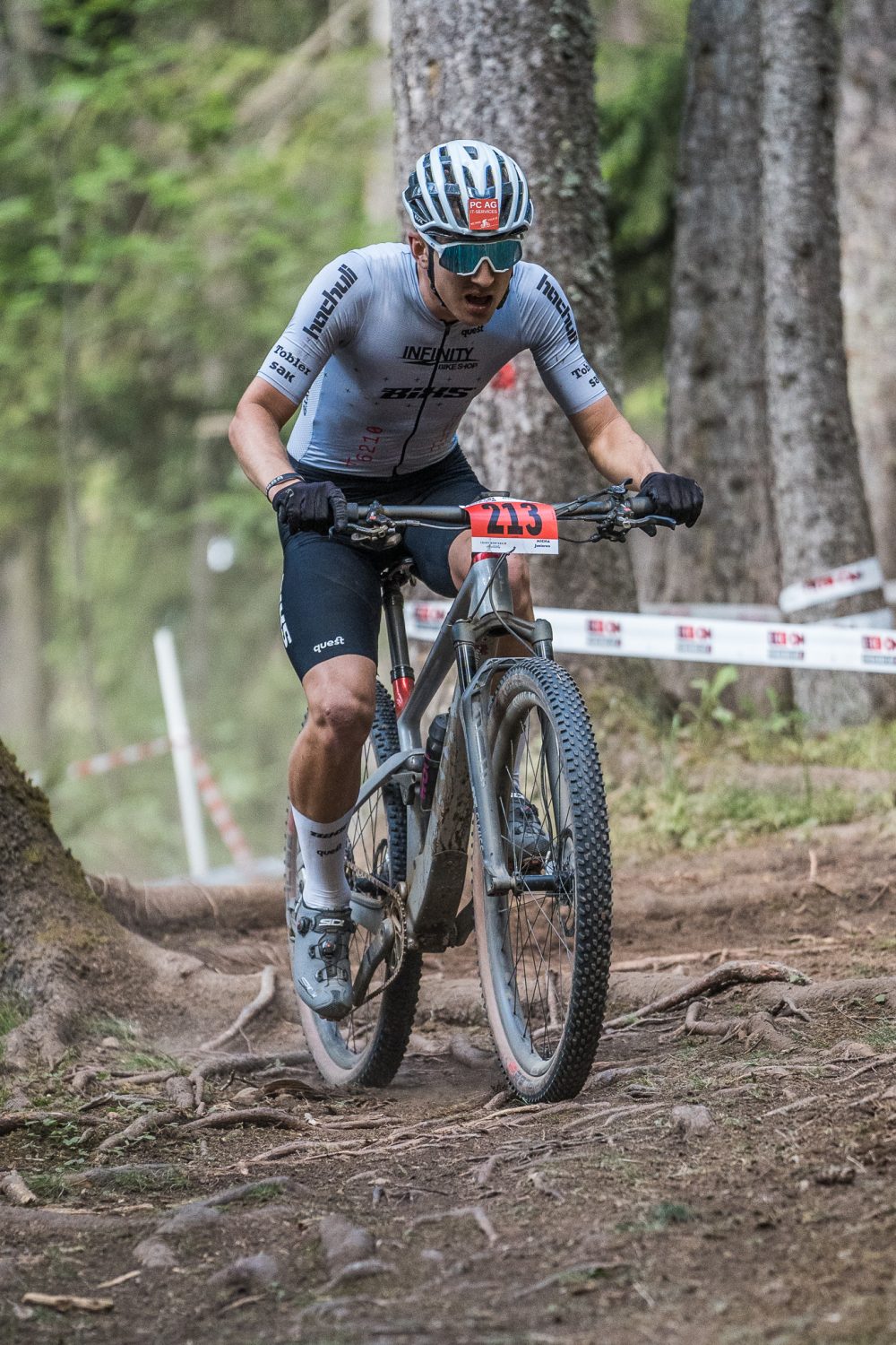 Ein Radfahrer mit Helm, Sonnenbrille und Renntrikot fährt mit einem E-Bike auf einem holprigen Waldweg, der mit Rennband und der Nummer 213 auf dem Lenker markiert ist.