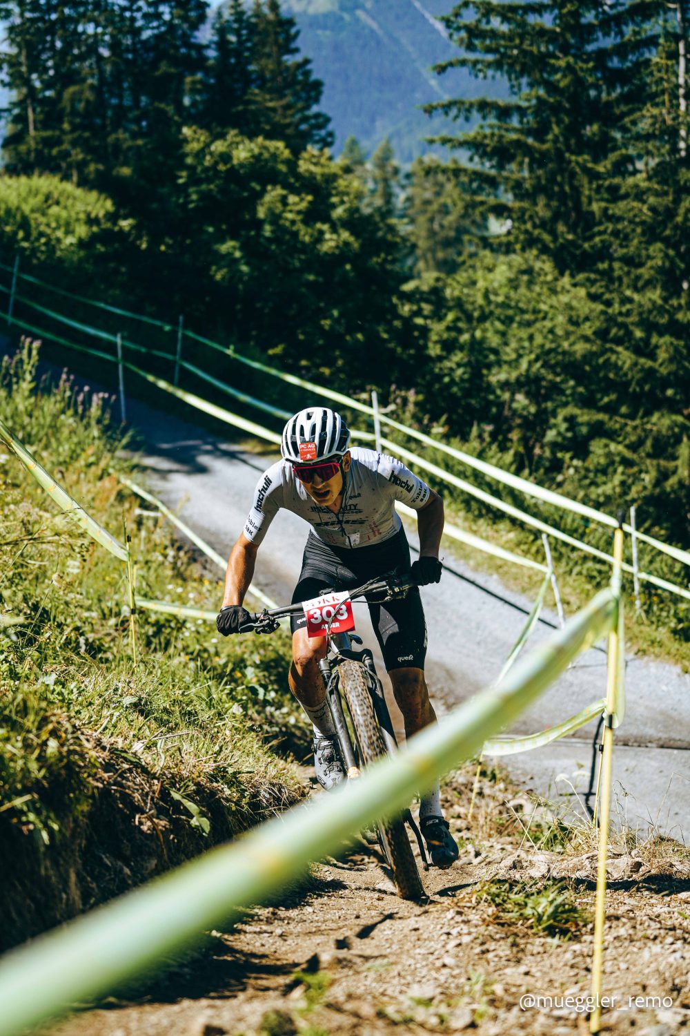Ein Mountainbiker mit Helm und Startnummer 306 rast an einem sonnigen Tag einen mit grünem Band markierten Feldweg hinunter, umgeben von üppigen Bäumen und Bergen.