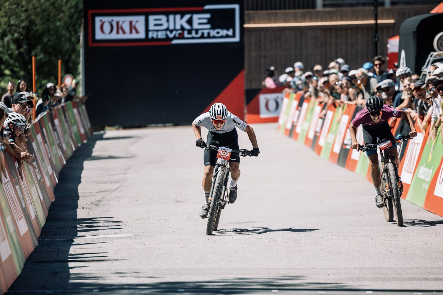 Zwei Radfahrer rasen in einem spannenden Radrennen auf die Ziellinie zu, angefeuert von den Zuschauern hinter den Absperrungen. Ein großes „Bike Revolution“-Banner sorgt für Spannung.