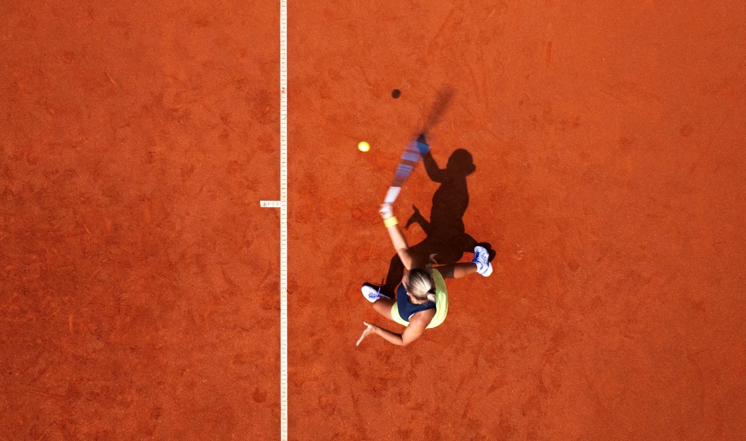 Eine Luftaufnahme eines Tennisspielers, der auf einem Sandplatz einen Ball schlägt. Er steht in der Nähe der weißen Mittellinie und sein Schatten ist auf dem Boden sichtbar.