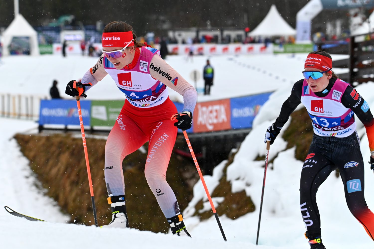 Zwei Athletinnen in Winterkleidung, darunter Ramona Schöpfer, bestreiten ein Langlaufrennen auf einer verschneiten Strecke. Eine Läuferin ist knapp vorn. Im Hintergrund sind Veranstaltungsbanner und Zuschauer zu sehen.