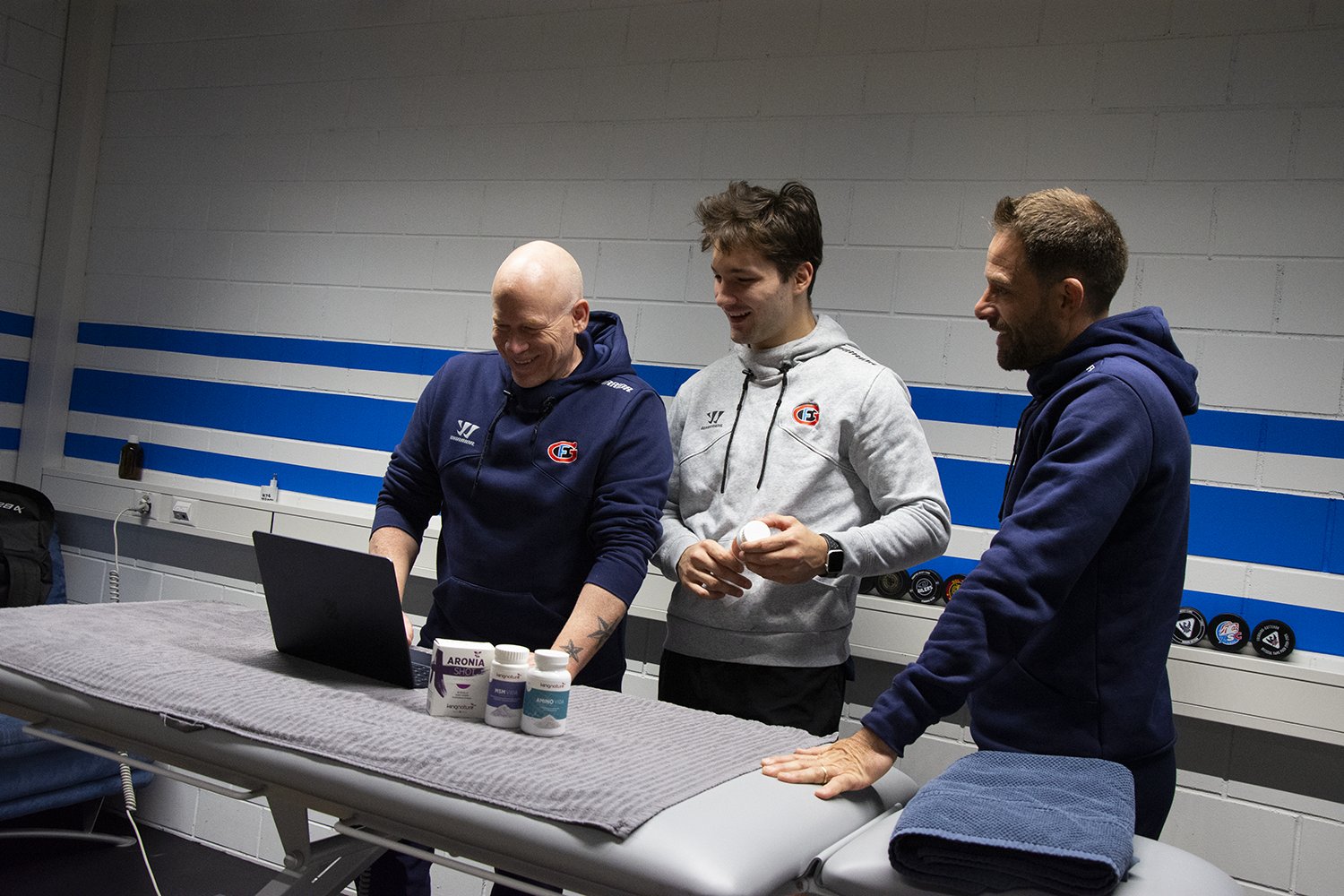 Drei Männer in Sportkleidung vom HC Fribourg-Gottéron aus der Nationalliga stehen lächelnd um einen Laptop auf einem Massagetisch herum in einem hell erleuchteten Raum mit blau-weißen Wänden. Auf dem Tisch stehen Flaschen und gefaltete Handtücher.