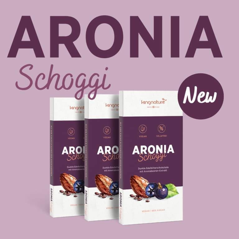 Drei Tafeln Aronia Schoggi stehen aufrecht vor einem hellrosa Hintergrund, mit der Aufschrift „ARONIA Schoggi“, dem Etikett „Neu“ und der Verpackung mit Abbildungen von Schokolade und Beeren. Jetzt lieferbar!.