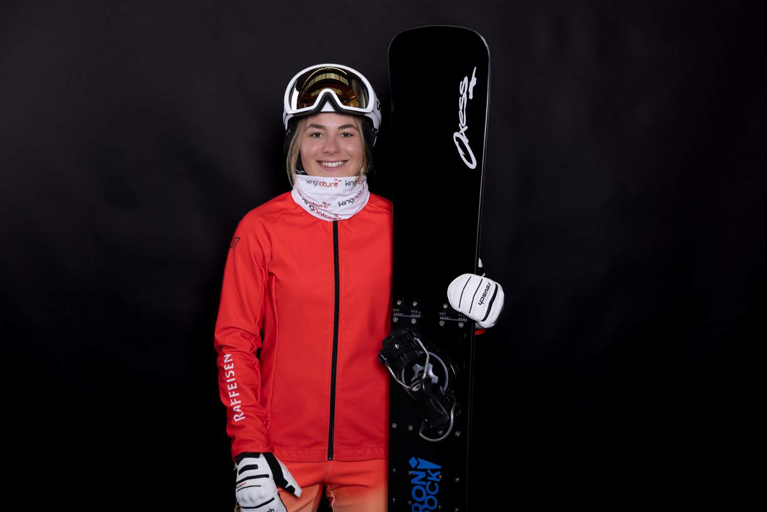 Ricarda Hauser, eine lächelnde Snowboarderin in roter Jacke und weißen Handschuhen, steht vor einem schwarzen Hintergrund und hält ihr Snowboard senkrecht. Sie trägt einen weißen Halswärmer, einen Helm und eine Skibrille auf dem Kopf.
