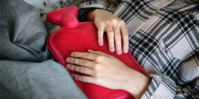 Eine Person in einem karierten Hemd legt sich mit einer roten Wärmflasche auf den Bauch und legt sanft ihre Hände darauf, um ihr erstaunliches Organ – den Magen – zu beruhigen.