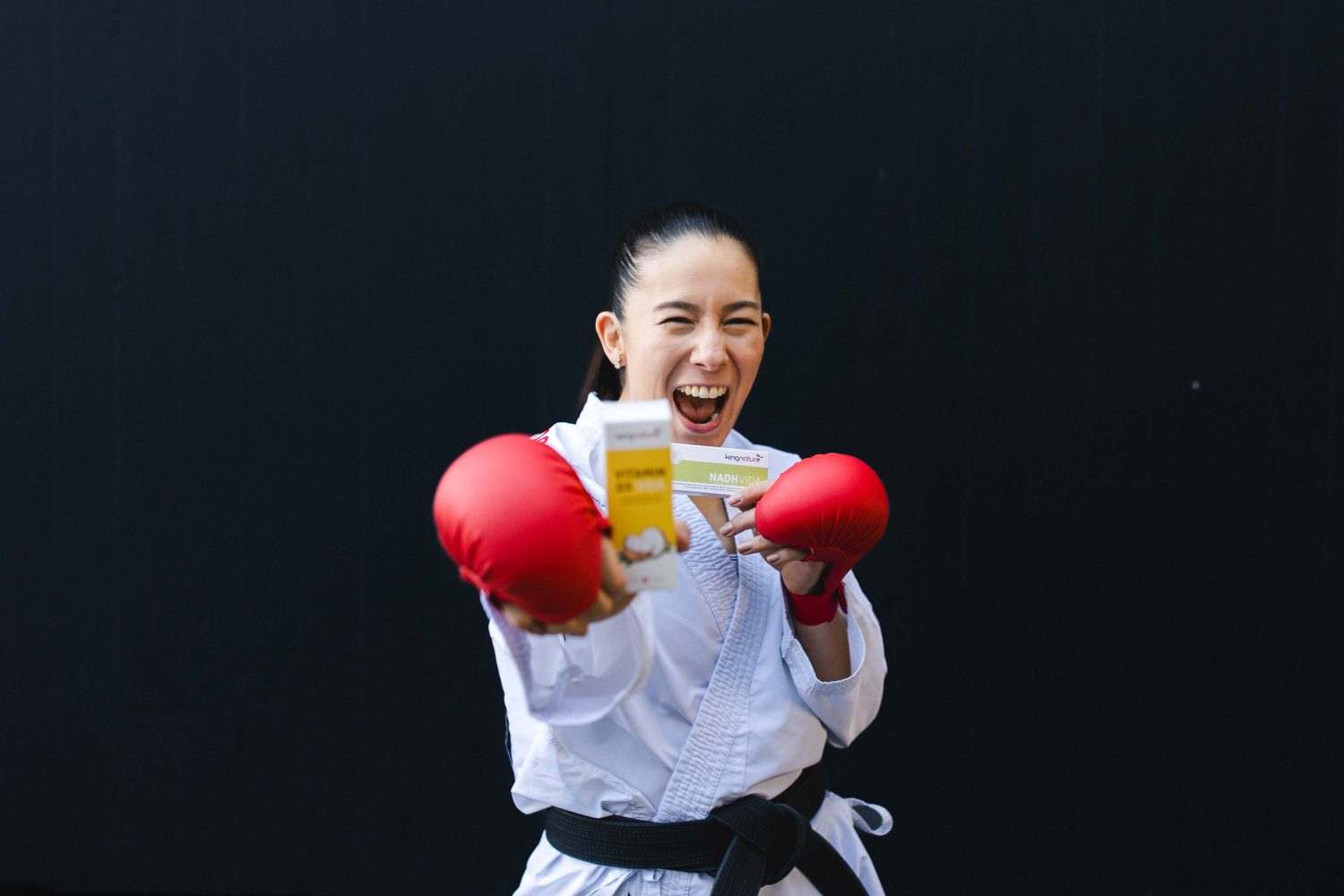 Maya Schärer, in einem Karateanzug und roten Handschuhen, lächelt energisch, während sie eine Schachtel hält und vor einem schlichten dunklen Hintergrund posiert.