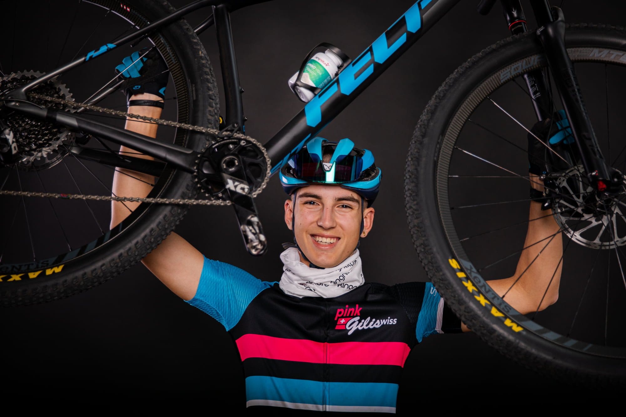 Ein lächelnder Radfahrer in farbenfrohem Rennoutfit und blauem Helm hebt vor dunklem Hintergrund stolz ein schwarzes Mountainbike über seinen Kopf und zeigt damit seine Leidenschaft fürs Radfahren.