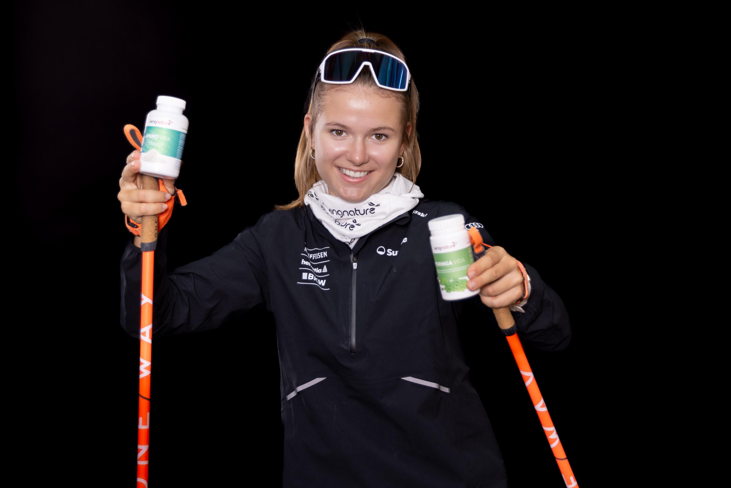 Ramona Schöpfer, lächelnd in einem schwarzen Sportjackett und mit Sonnenbrille, hält zwei orangefarbene Skistöcke und zwei Flaschen mit Nahrungsergänzungsmitteln in der Hand und posiert vor einem schwarzen Hintergrund.