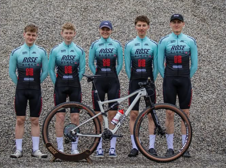 Fünf junge Radfahrer in passenden blau-schwarzen Uniformen des ROSE Bikes Factory Racing Teams stehen im Freien in einer Reihe und posieren mit einem Mountainbike vor einem Schotterhintergrund.